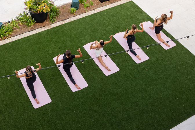 Aerial view of five yoga participants on pink mats doing a low-lunge twist on a green turf lawn under string lights — outdoor group yoga/fitness class.