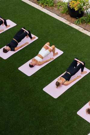 Outdoor fitness class with women on pink mats doing glute bridge exercises using pink resistance bands on a green lawn beside landscaped planters