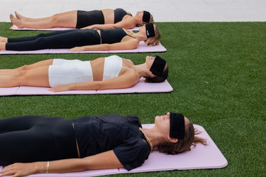 Group of women reclining on pink yoga mats on green turf outdoors, blindfolded and relaxed during a guided meditation or restorative yoga session.