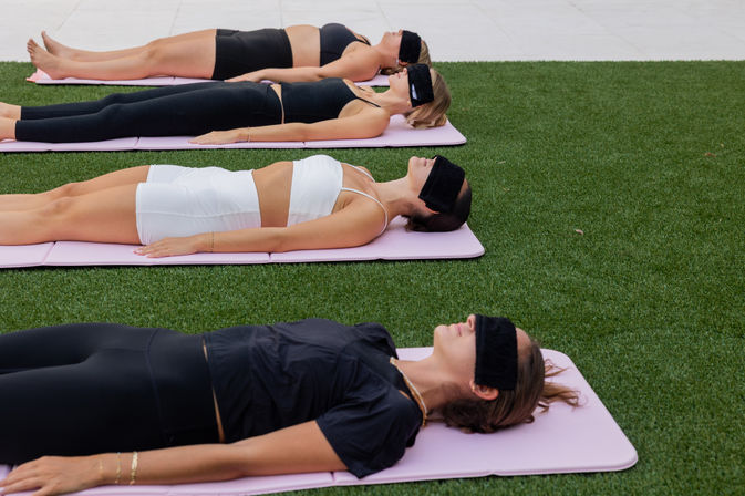 Group of women reclining on pink yoga mats on green turf outdoors, blindfolded and relaxed during a guided meditation or restorative yoga session.
