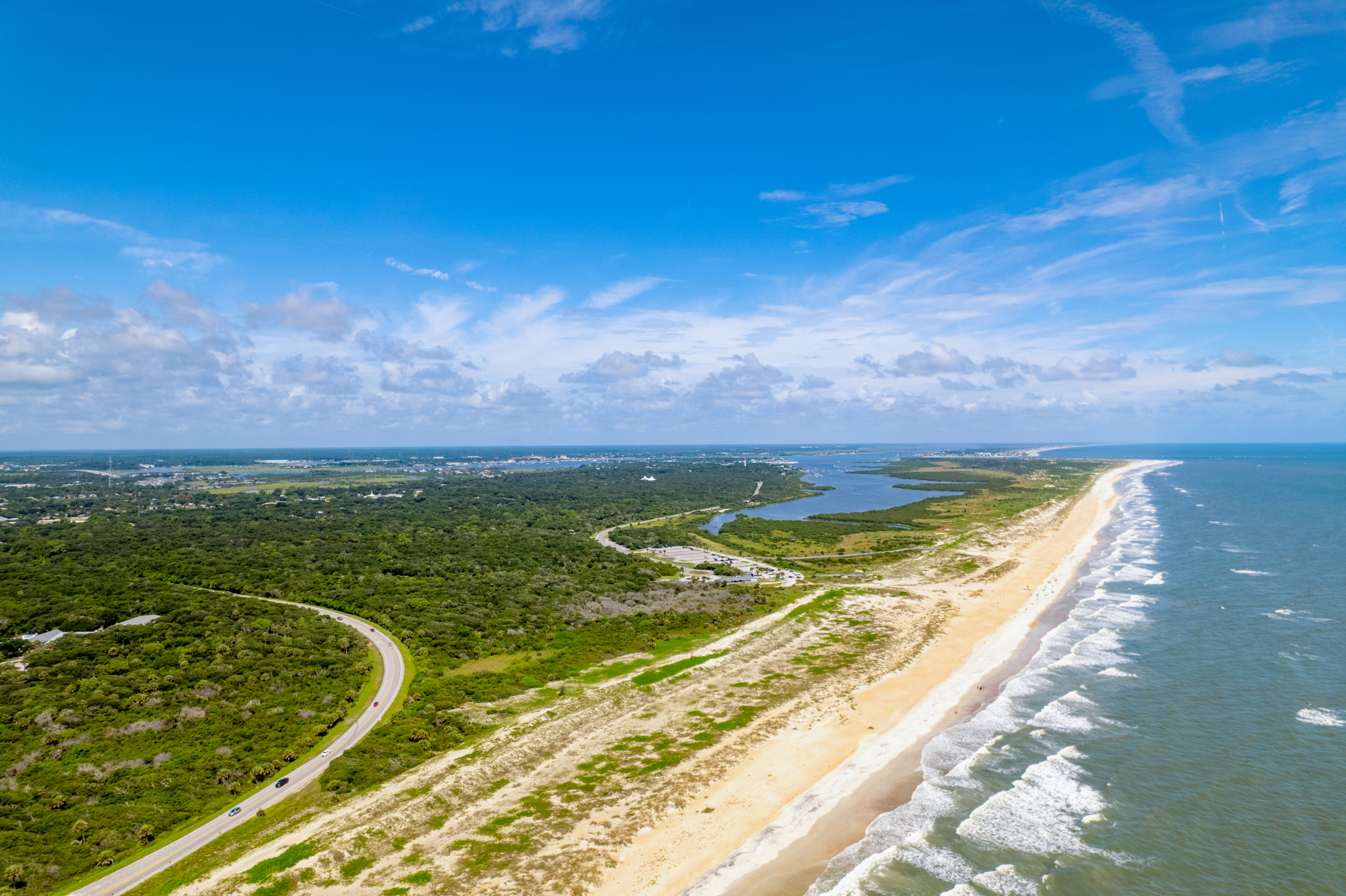 Sunny aerial view of a long sandy beach and surf along a barrier island with coastal marshes, a winding road, and bright blue sky
