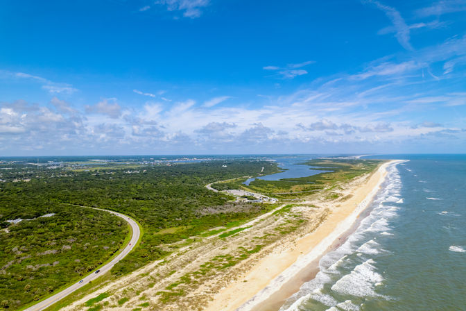 Sunny aerial view of a long sandy beach and surf along a barrier island with coastal marshes, a winding road, and bright blue sky
