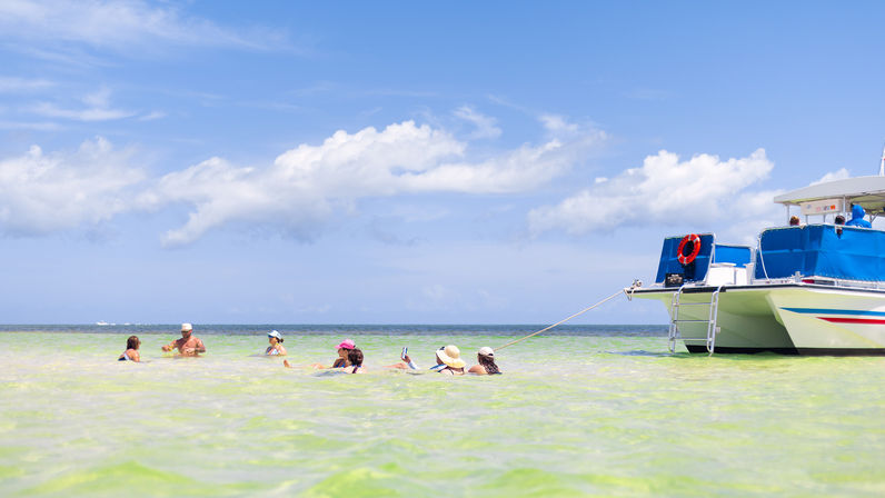 Vacationers wading in shallow turquoise tropical lagoon near an anchored white-and-blue catamaran under a sunny blue sky with scattered clouds