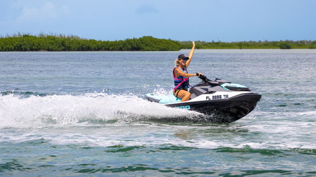 Woman in life vest waving while riding a turquoise jet ski across calm Florida coastal waters with mangroves in the distance