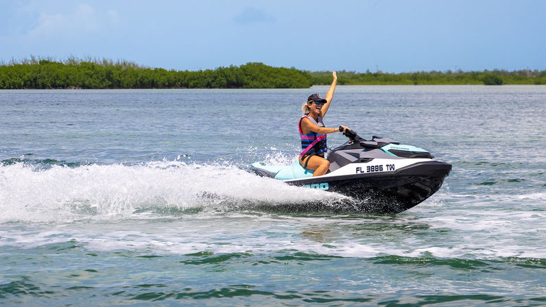 Woman in life vest waving while riding a turquoise jet ski across calm Florida coastal waters with mangroves in the distance