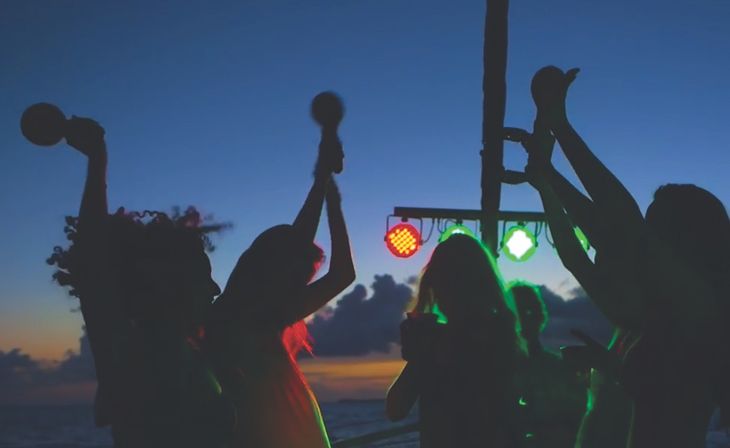 Silhouetted group at a sunset beach party, dancing and raising maracas under red and green lights with the ocean and colorful sky behind them.