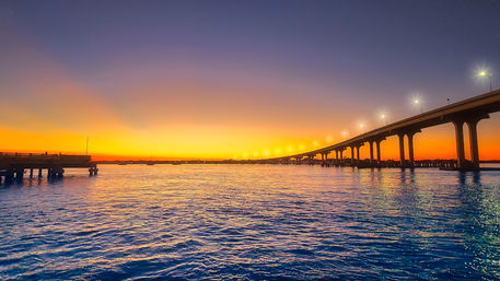 Golden-orange sunset over a coastal bay with a long illuminated bridge and wooden pier reflecting on rippled blue water.
