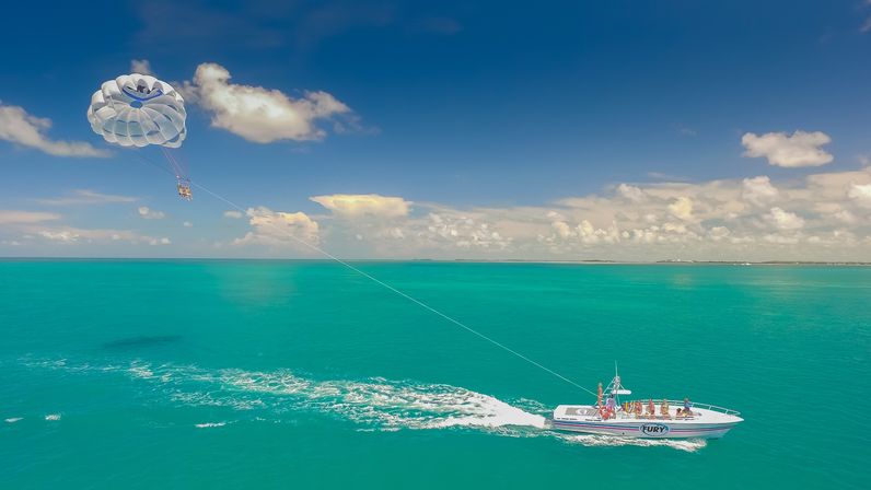 Aerial view of a white speedboat towing a parasail with riders over turquoise tropical ocean under a bright blue sky with fluffy clouds.