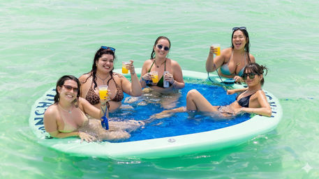 Five women in bikinis relaxing on a round inflatable float in clear turquoise water, holding yellow drinks and smiling during a sunny summer beach day.