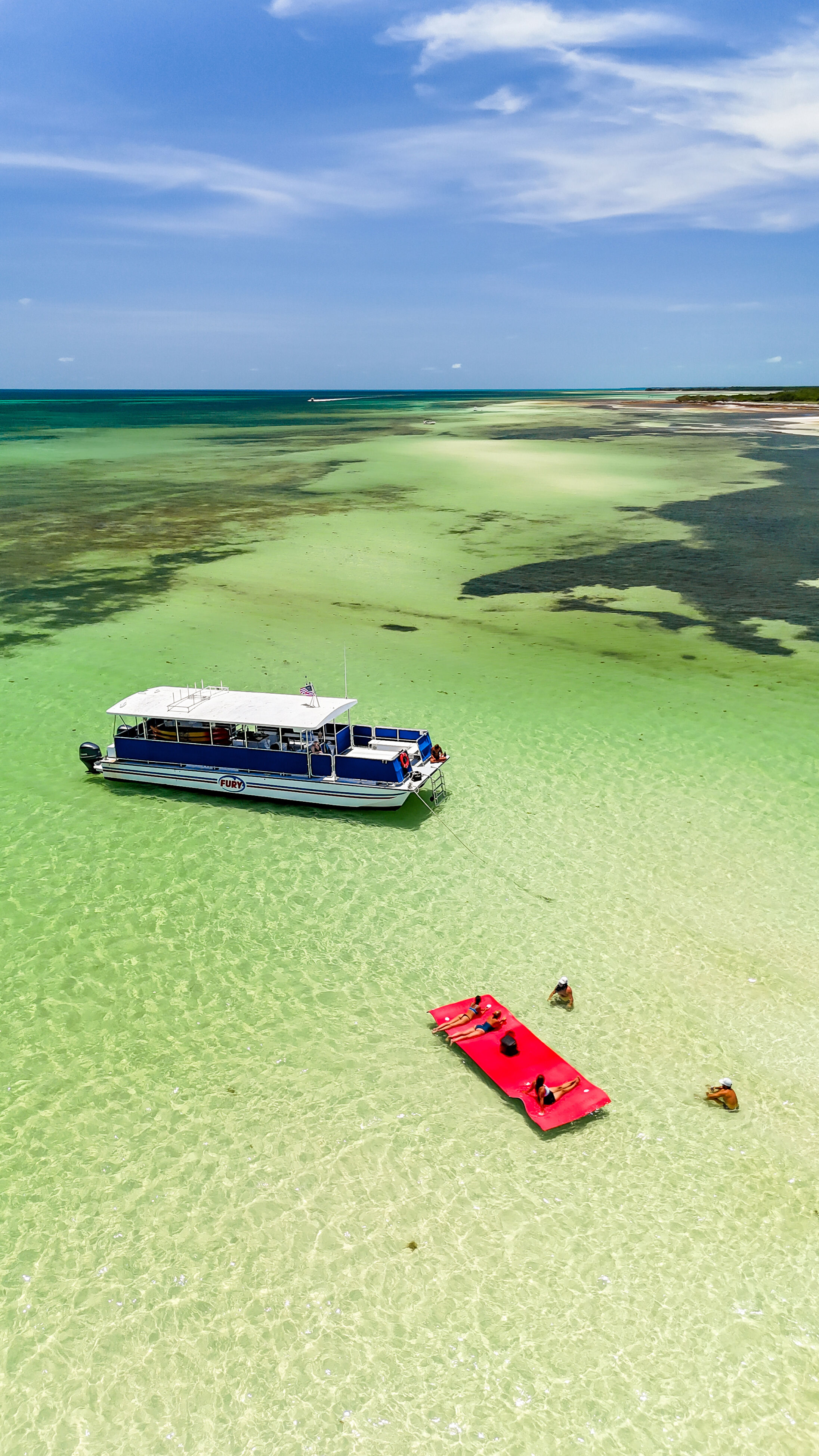 Aerial drone shot of tropical shallow turquoise water and sandbars with an anchored blue tour boat, a bright red floating mat with sunbathers, and swimmers wading under a sunny sky.