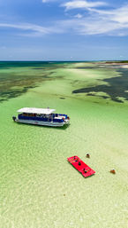 Aerial drone shot of tropical shallow turquoise water and sandbars with an anchored blue tour boat, a bright red floating mat with sunbathers, and swimmers wading under a sunny sky.