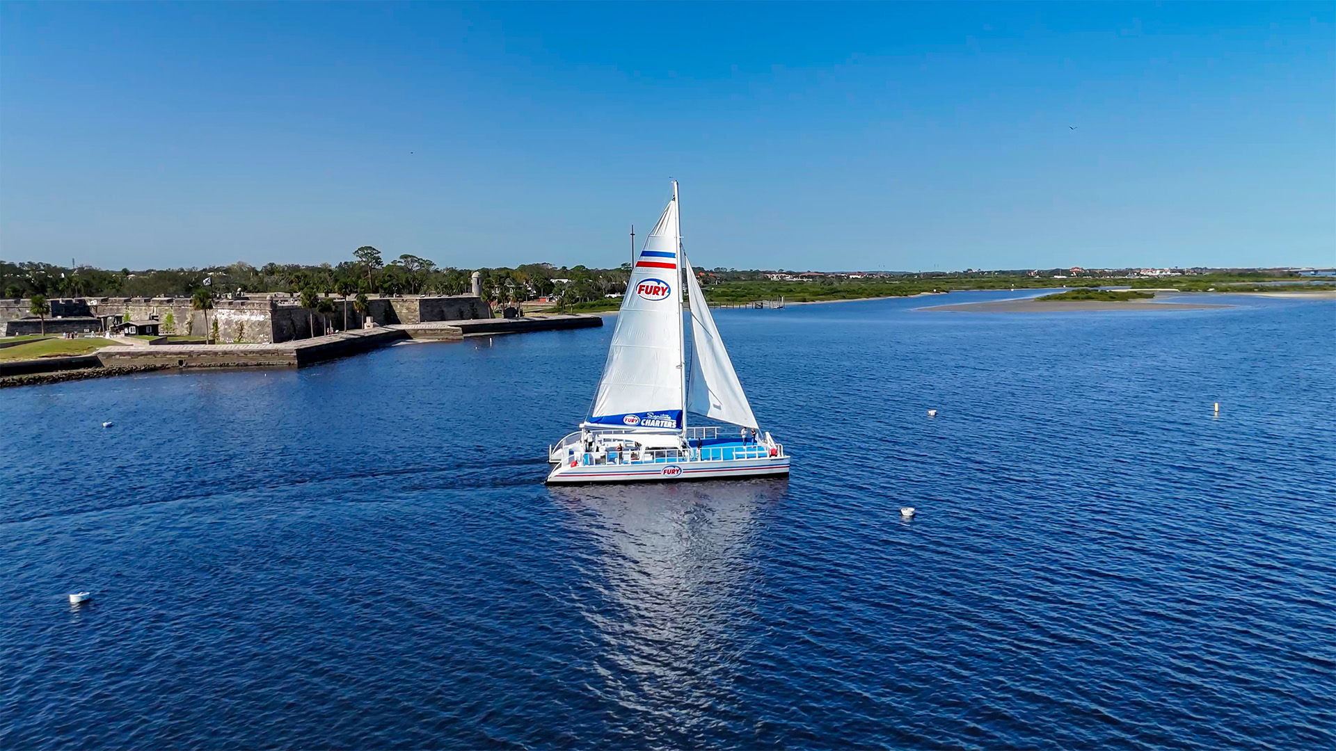White catamaran sailboat gliding across a calm blue bay past a low stone coastal fort and grassy shoreline under a clear sunny sky