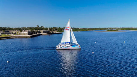 White catamaran sailboat gliding across a calm blue bay past a low stone coastal fort and grassy shoreline under a clear sunny sky