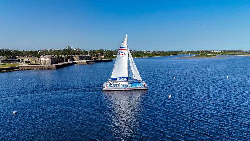 White catamaran sailboat gliding across a calm blue bay past a low stone coastal fort and grassy shoreline under a clear sunny sky