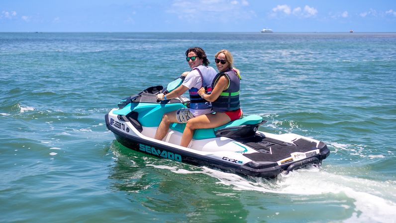 Two people in life vests smiling as they ride a turquoise-and-white jet ski together on clear blue coastal waters under a sunny sky