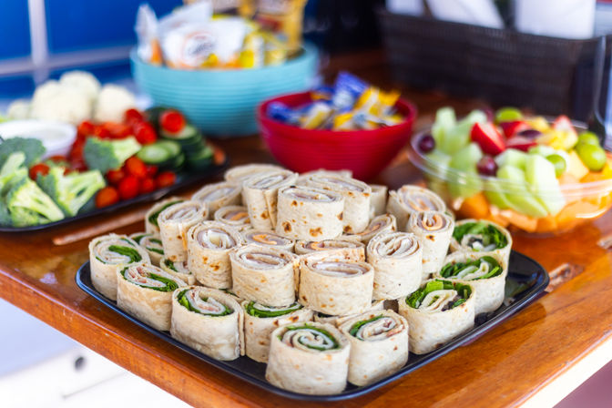 Plate of tortilla wrap pinwheel sandwiches stacked on a wooden table with a fresh vegetable platter, fruit bowl and snack bowls — colorful catered appetizer spread for a party or buffet