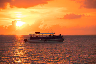 Silhouetted passenger boat on calm ocean at a fiery orange sunset, sun sinking behind clouds with golden reflections on rippling water.