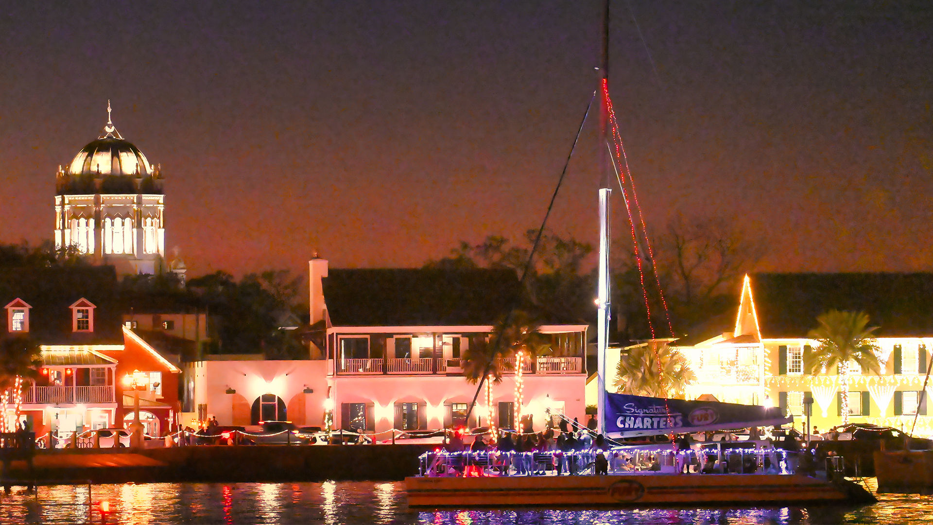 Festive nighttime waterfront scene with a glowing domed landmark, illuminated shops along the marina, and a sailboat strung with colorful lights reflecting on the water.