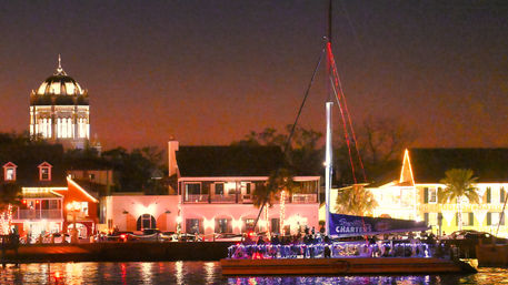 Festive nighttime waterfront scene with a glowing domed landmark, illuminated shops along the marina, and a sailboat strung with colorful lights reflecting on the water.