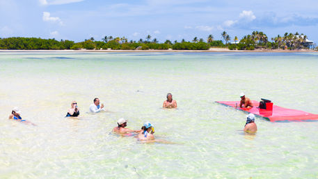 Group of people wading and relaxing in shallow turquoise lagoon with a palm‑lined tropical island and sunny sky in the background.