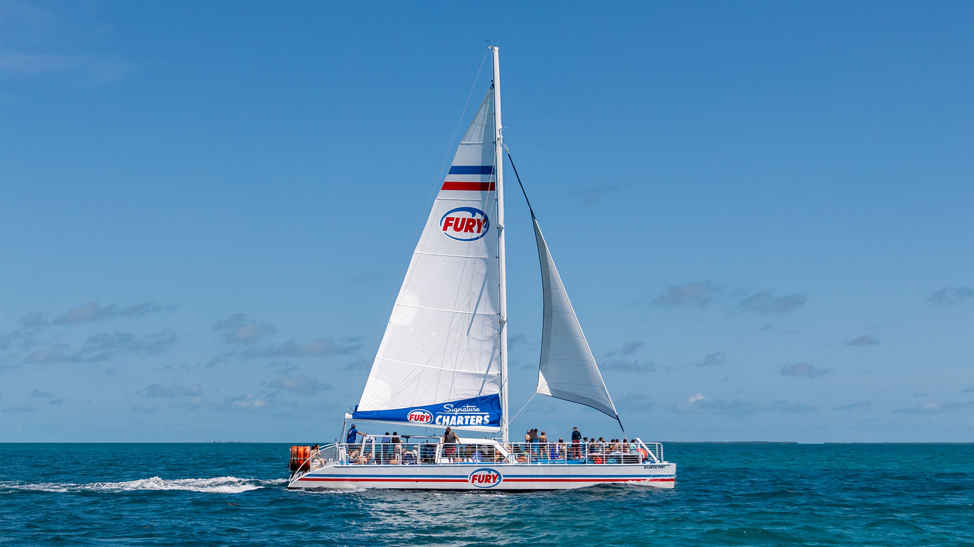 Sunlit white catamaran charter with tall sails carrying passengers across turquoise ocean under a clear blue sky