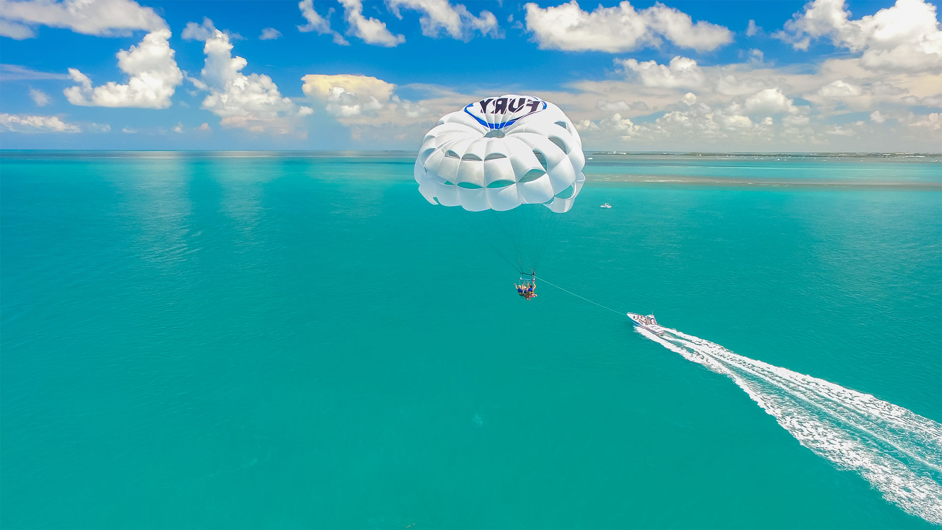Parasailing over turquoise tropical ocean with a white parasail and riders towed by a speedboat under a bright blue sky dotted with fluffy clouds and a distant shoreline