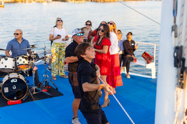 People enjoying a lively boat cruise on a blue deck at sunset — two men pulling a rope, a small live band with drums, and guests in colorful outfits watching with water and shoreline in the background.