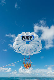 Three parasailers suspended under a white round parasail with blue accents above turquoise ocean, sunny blue sky and fluffy clouds — tropical coastal water sports scene.
