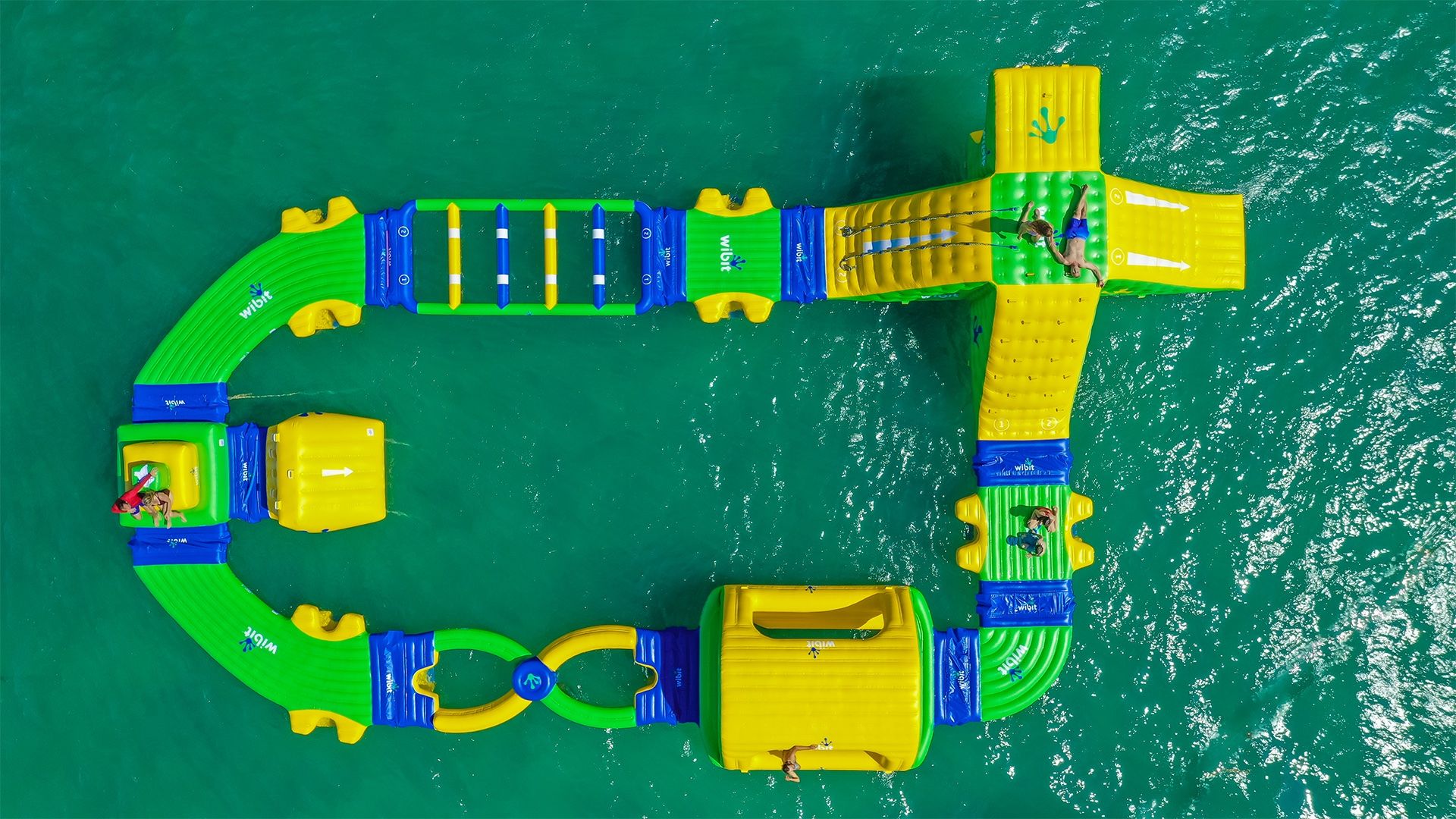 Aerial view of a green-and-yellow inflatable water park obstacle course floating on turquoise ocean with people climbing, sliding, and swimming — vibrant summer activity on the water.