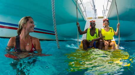 Three smiling adults enjoying turquoise tropical water by a catamaran: one woman swimming and two people in yellow life vests sitting on the boat ladder with snorkel masks, vacation snorkeling.