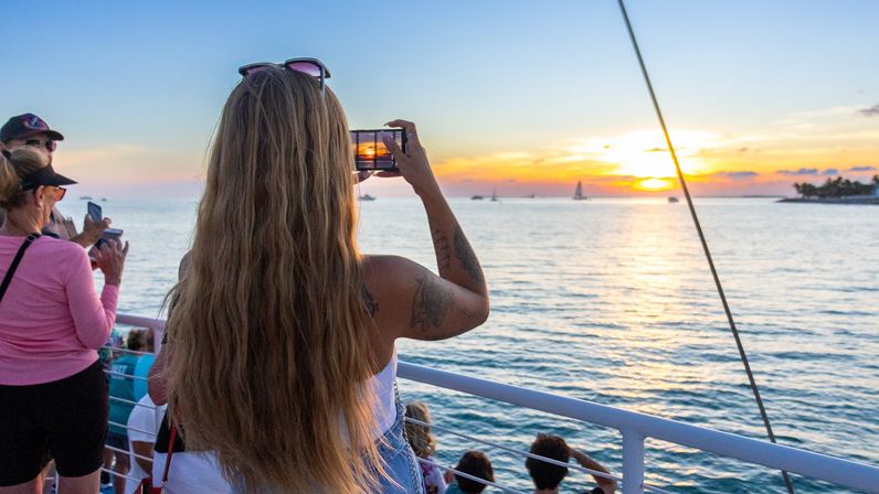 Person with long hair snapping a photo of a vibrant golden sunset over the calm ocean from a crowded boat deck with sailboats on the horizon.