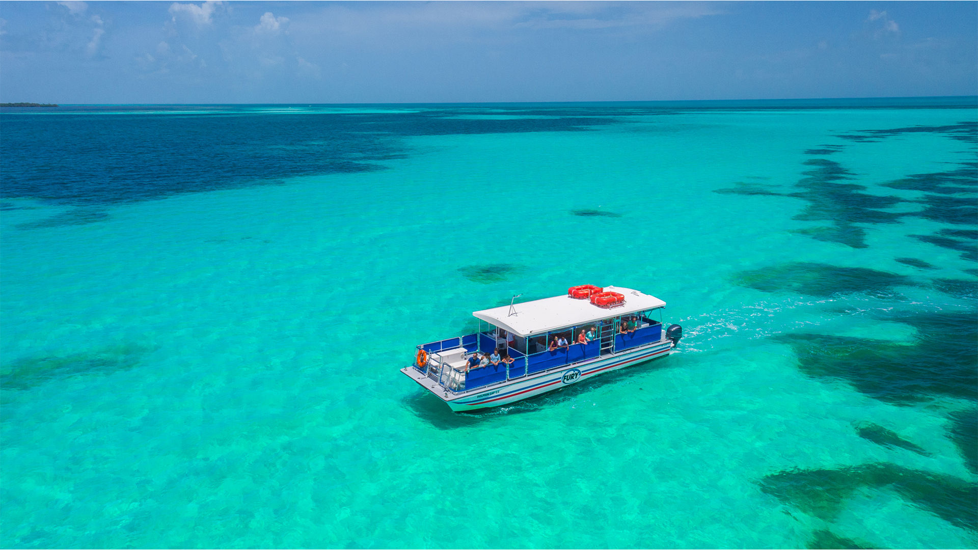Bright tour boat gliding over crystal-clear turquoise tropical waters dotted with dark coral patches under a sunny blue sky