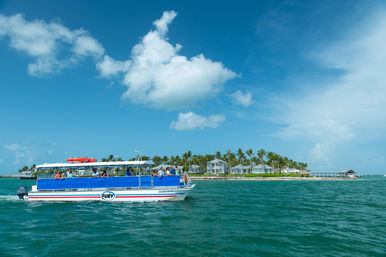 Open-air tour boat cruising turquoise waters past a palm-lined tropical island with beachside bungalows under a bright blue sky.