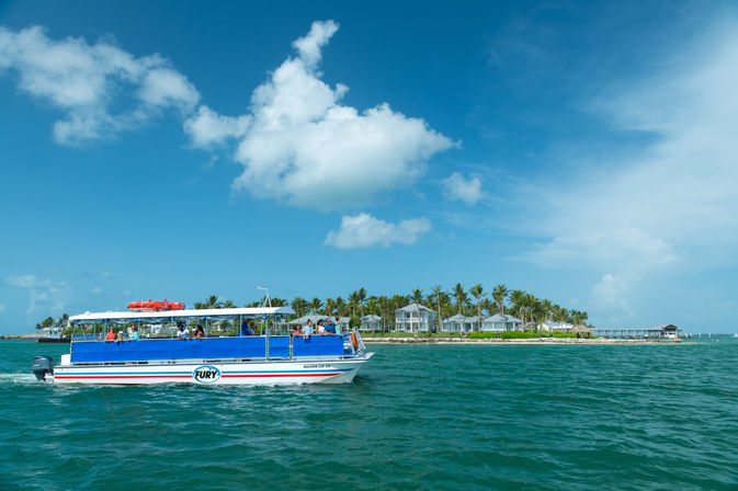 Open-air tour boat cruising turquoise waters past a palm-lined tropical island with beachside bungalows under a bright blue sky.
