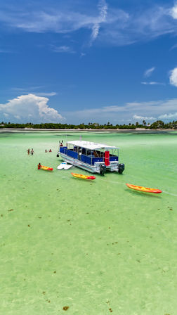 White-and-blue tour boat anchored in shallow turquoise lagoon with orange kayaks and people wading near a palm-lined sandy island under a bright blue sky.