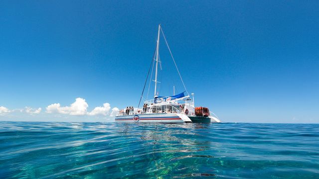 White catamaran tour boat with tall mast and passengers floating on crystal-clear turquoise ocean under a bright blue sky with a few fluffy clouds.