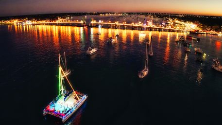 Aerial view of a brightly lit party boat with green and multicolored lights among anchored sailboats, reflections shimmering on calm harbor waters at dusk with an illuminated pier and city lights along the shoreline.