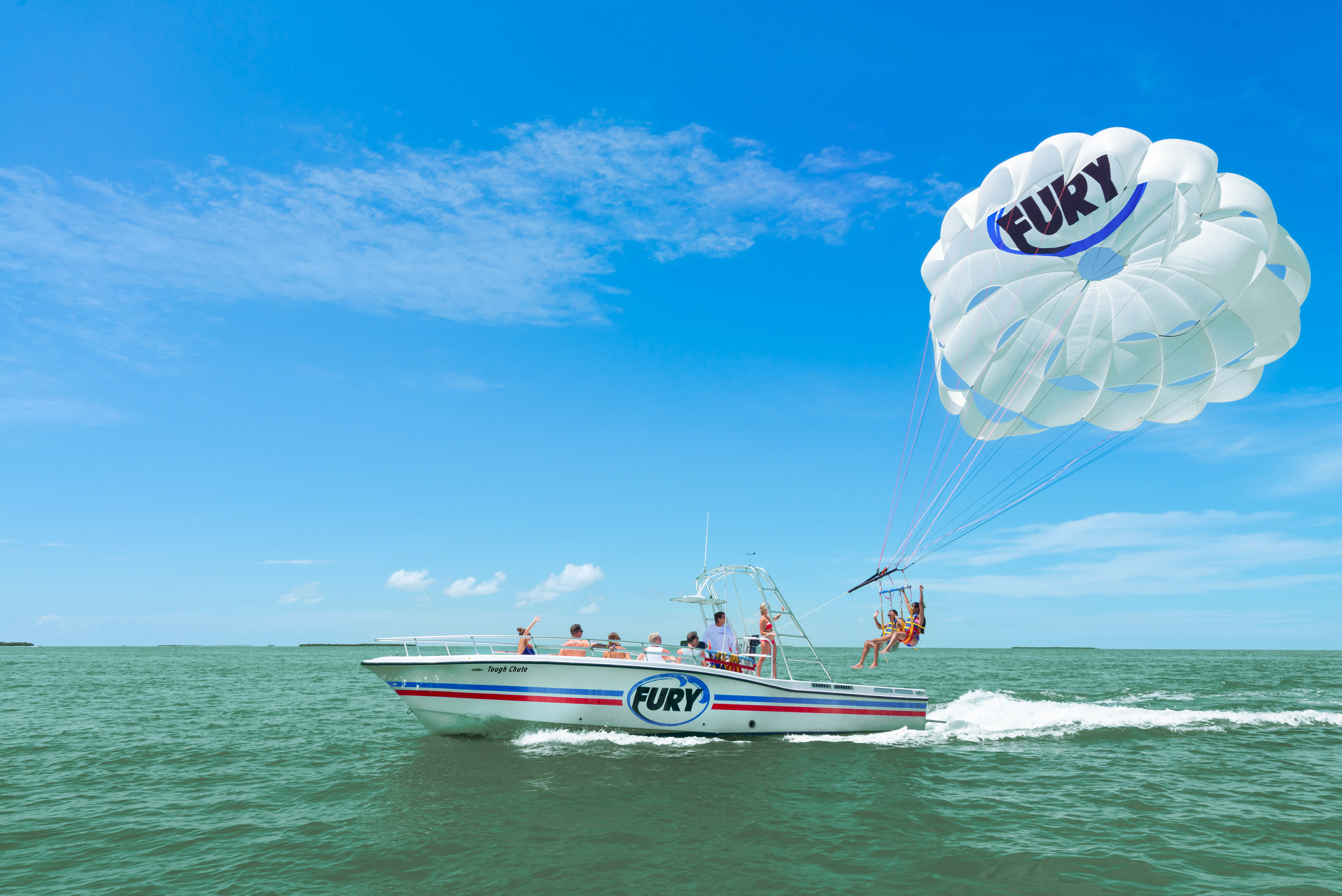 Thrilling parasailing scene: speedboat with passengers towing two people beneath a large white parasail over turquoise coastal waters under a clear blue sky.