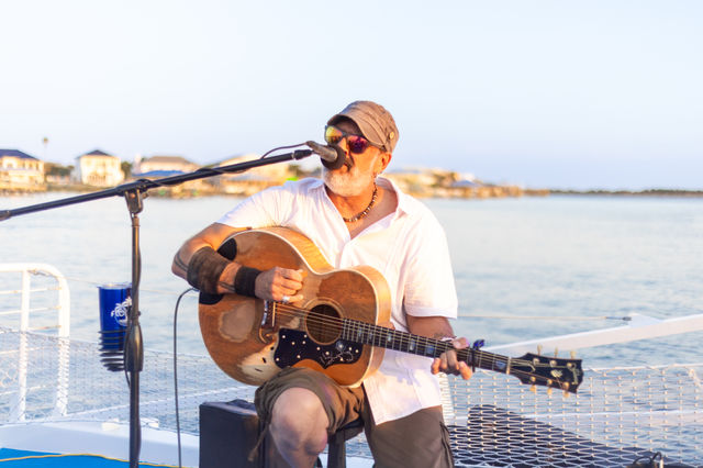 Bearded musician in sunglasses and a cap playing a worn acoustic guitar and singing into a microphone on a boat deck by the waterfront at sunset, shoreline houses in the background.