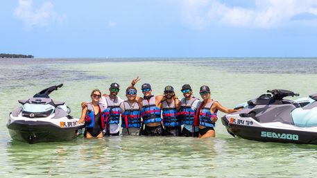 Seven people in colorful life jackets standing waist-deep in clear turquoise water between two Sea-Doo jet skis, smiling and posing under a sunny sky near a tropical shoreline.