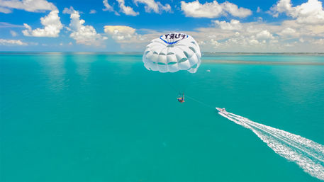 Parasailers suspended beneath a white parasail, towed by a speedboat across turquoise tropical ocean under a bright blue sky with fluffy clouds.