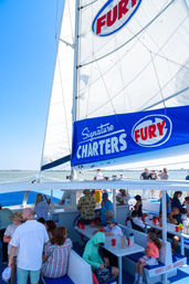 Families and friends on a sunny catamaran day cruise beneath a tall white sail with a blue branded banner, calm coastal waters and picnic-style seating on deck.