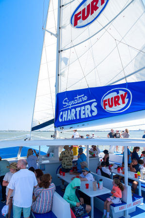 Families and friends on a sunny catamaran day cruise beneath a tall white sail with a blue branded banner, calm coastal waters and picnic-style seating on deck.