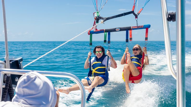 Two people parasailing in tandem above clear turquoise ocean, wearing life vests and sunglasses, suspended from colorful harnesses and towed by a speedboat on a sunny blue-sky day