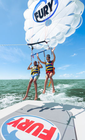 Two people tandem parasailing above turquoise ocean on a sunny day, wearing yellow life jackets suspended from a white parasail with blue accents, boat deck visible in the foreground.