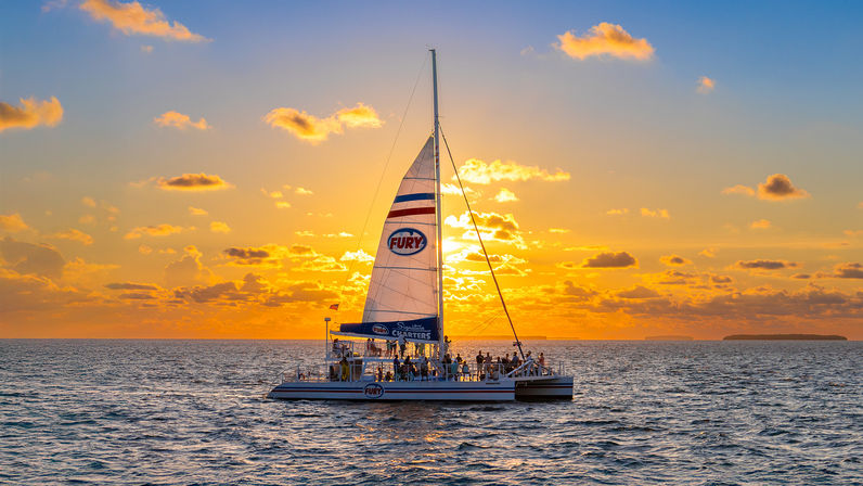 Catamaran sunset cruise with passengers silhouetted against a glowing golden-orange sky, scattered clouds and calm ocean waters on the horizon.