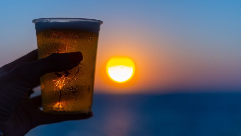 Hand holding a cold plastic cup of beer with condensation, silhouetted against a golden-orange ocean sunset over the horizon.