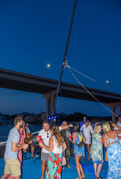 Evening boat party: people dancing with red cups and live music on deck beneath a lit highway bridge by waterfront homes.