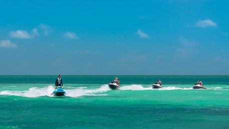 Four riders on jet skis moving in a line across a turquoise tropical sea with white spray and a clear blue sky — beach water sports scene.