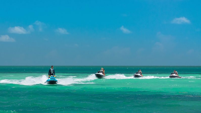Four riders on jet skis moving in a line across a turquoise tropical sea with white spray and a clear blue sky — beach water sports scene.